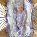 Newborn baby in purple ruffled knitted romper and headband lying in a bassinet