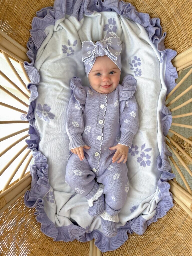 Newborn baby in purple ruffled knitted romper and headband lying in a bassinet
