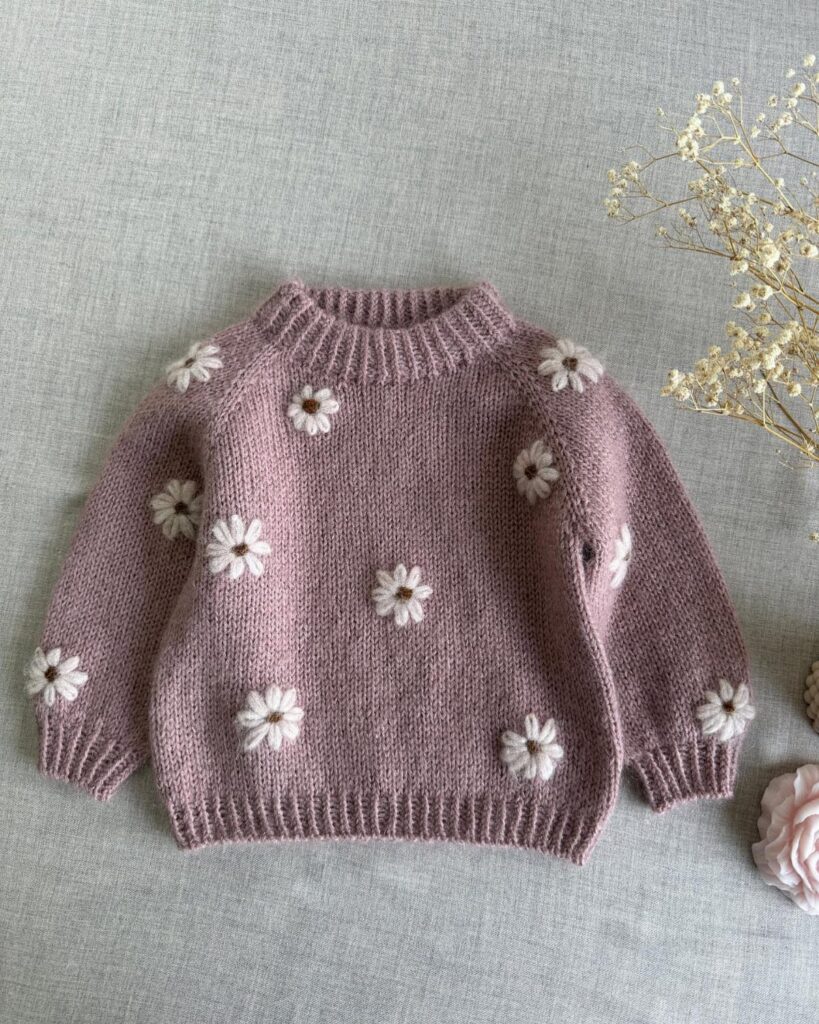 Mauve knitted baby sweater with white daisy flowers on a grey background