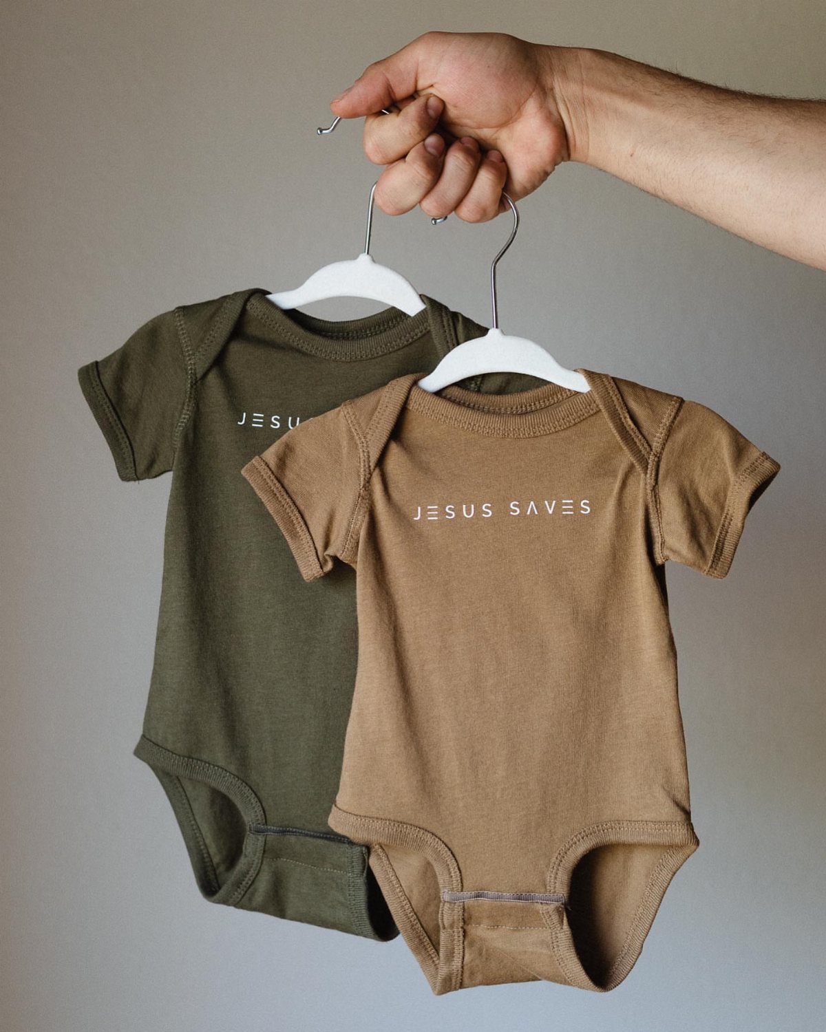 Two baby bodysuits in olive and tan with Jesus Saves printed on them.
