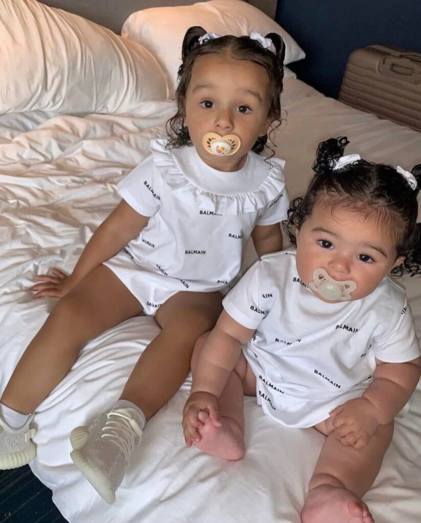Two young children wearing matching white Balmain designer outfits sitting on a bed