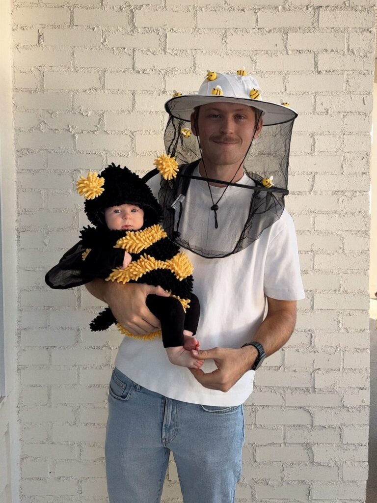 Man in beekeeper hat holding a baby wearing a bumblebee costume against white brick.