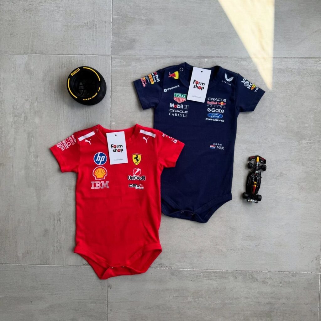 Red and navy blue baby bodysuits with racing logos on a grey floor