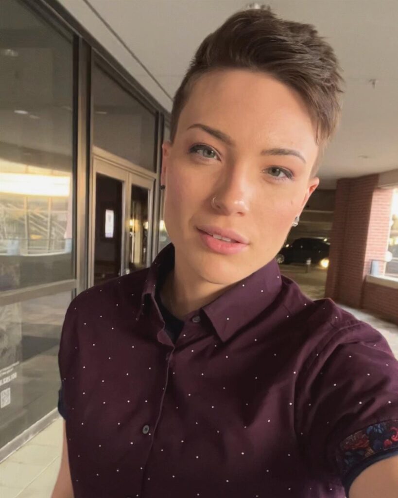 Person with short hair wearing a maroon dotted button-up shirt in a hallway.