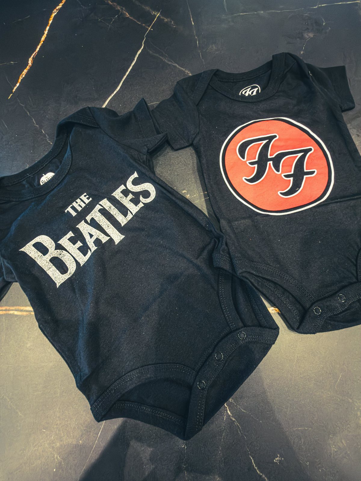 Two black infant bodysuits with Beatles and FF logos on a dark surface.