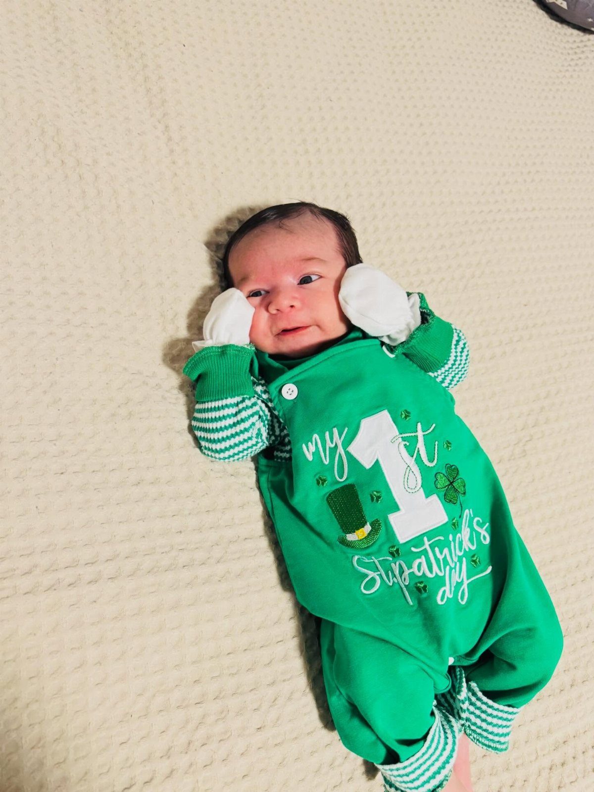 Newborn baby wearing a green St Patrick's Day outfit lying on a cream blanket.