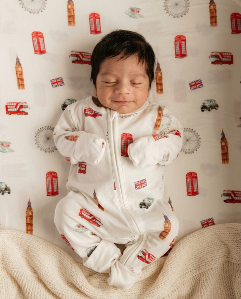 Sleeping newborn baby wearing white London-themed patterned pajamas on matching bedding.