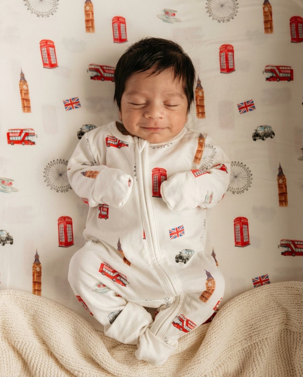 Sleeping newborn baby wearing white London-themed patterned pajamas on matching bedding.