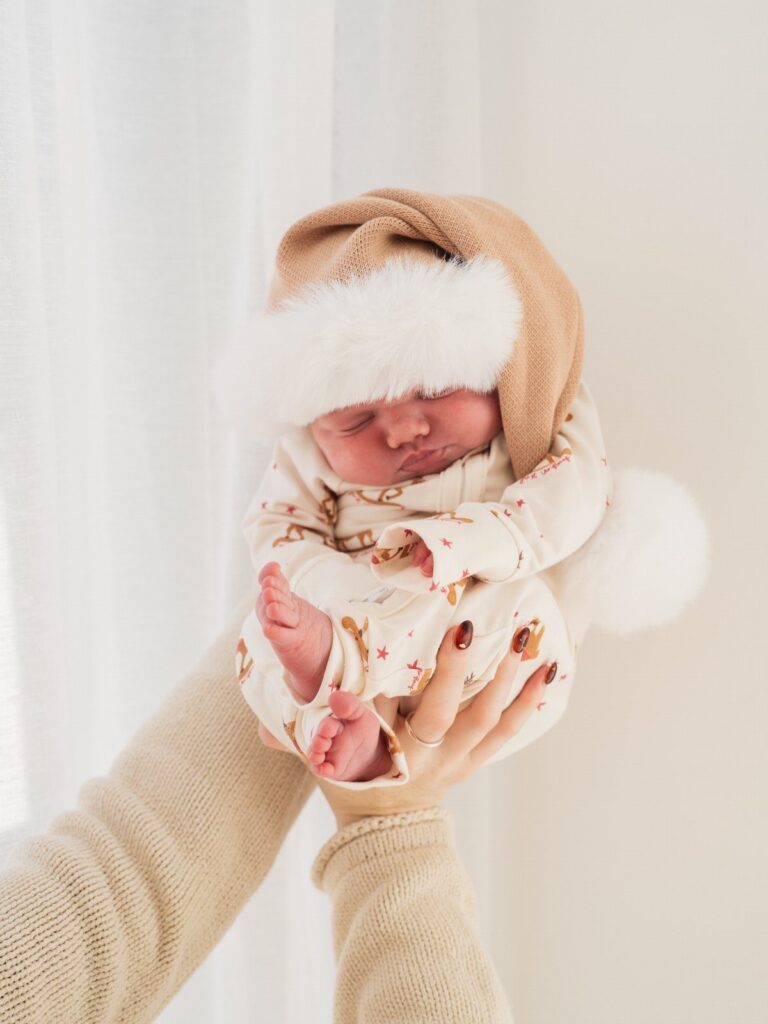 Sleeping newborn baby wearing a tan Christmas hat and white patterned onesie.
