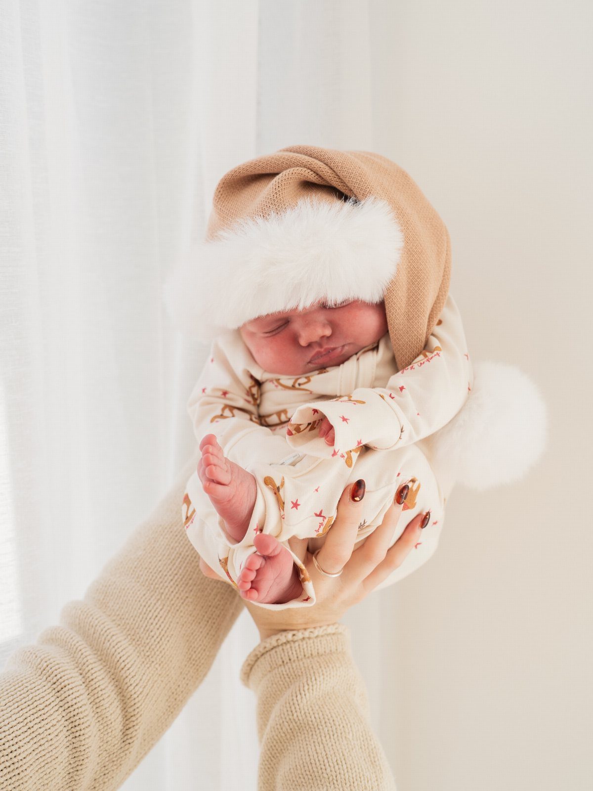 Sleeping newborn baby wearing a tan Christmas hat and white patterned onesie.