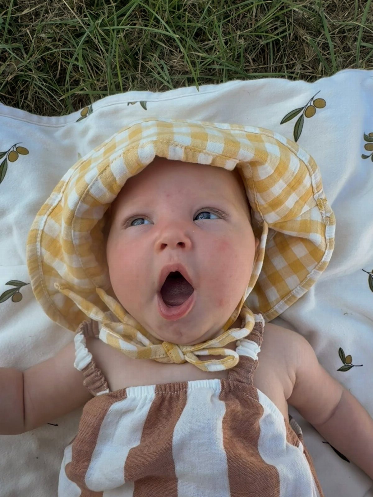 Newborn baby wearing a yellow checkered sun hat and striped romper on grass